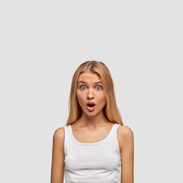 Stunned blonde young woman in casual white vest, hears horror news or something bad, isolated over white studio wall