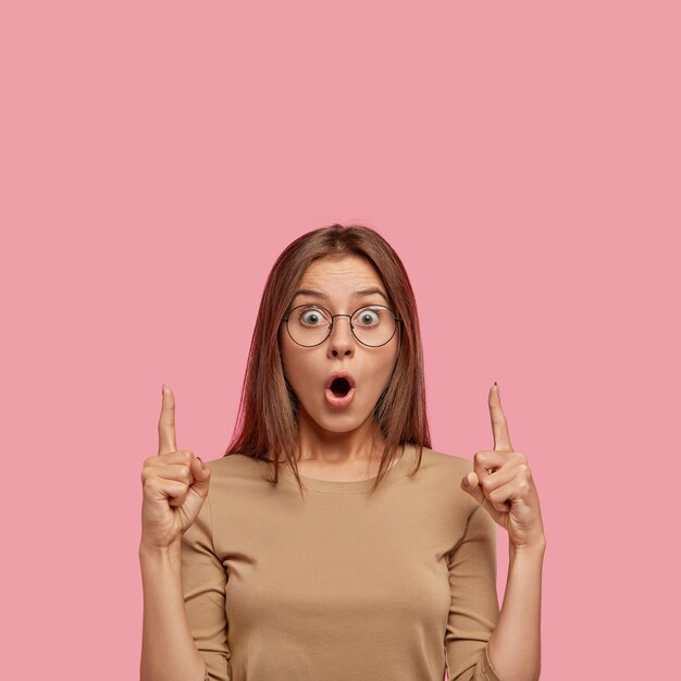 Stunned Caucasian female has shocked expression, points upwards with both index fingers, cant believe in something, shows blank space above head, has appealing appearance. People and reaction