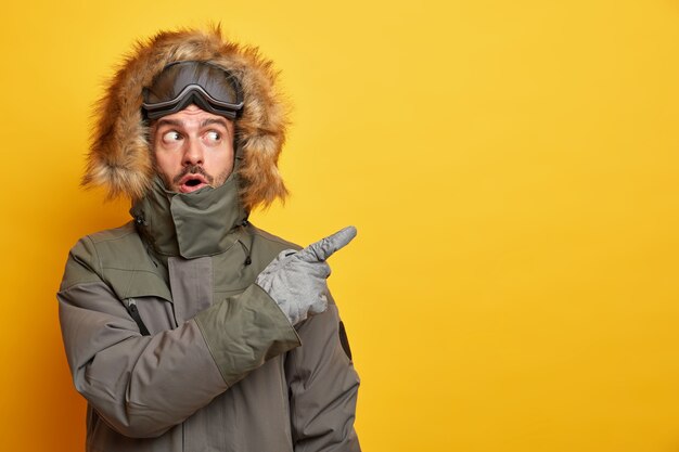 Stunned Caucasian man in winter clothes points away on blank space with surprised expression goes skiing during cold day wears jacket and gloves enjoys chilly weather.