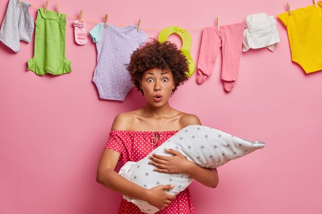 Stunned curly haired mother holds infant baby, finds out about childs serious illness, keeps mouth widely opened, poses 