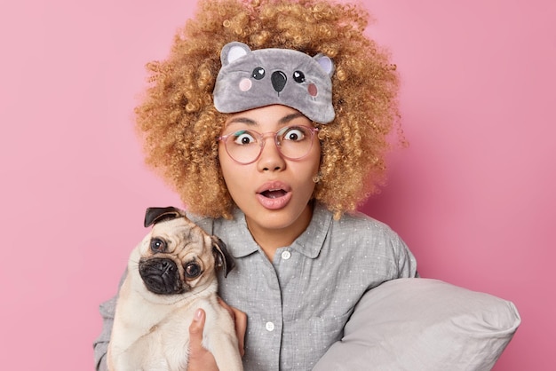 Stunned curly woman stares bugged eyes and opened mouth cannot believe in shocking news holds pug dog and soft pillow dressed in nightwear poses indoor People animals and sleeping time concept