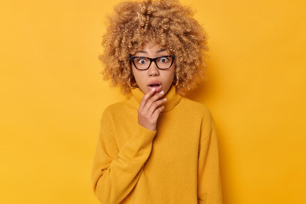 Stunned curly young woman gasps from wonder holds breath stares at something horrible keeps jaw dropped dressed in casual jumper isolated over vivid yellow background. Human reactions concept