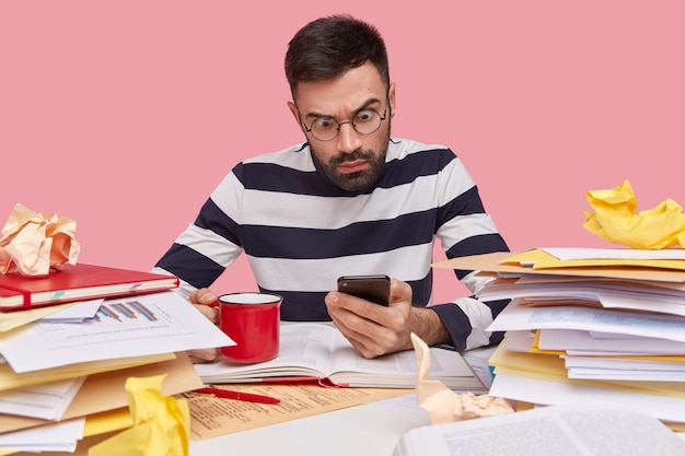 Stordito emotivo giovane uomo con la barba lunga tiene moderno cellulare, indossa occhiali rotondi trasparenti e maglione a strisce, beve caffè caldo aromatico