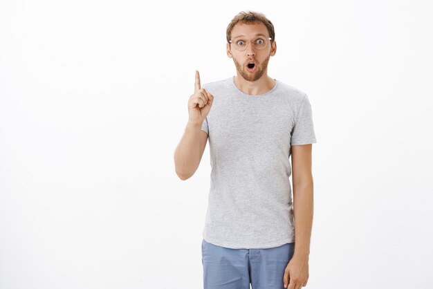 Stunned excited charming mature guy with bristle gasping opening mouth in surprise while making up great plan raising index finger in eureka gesture over white wall