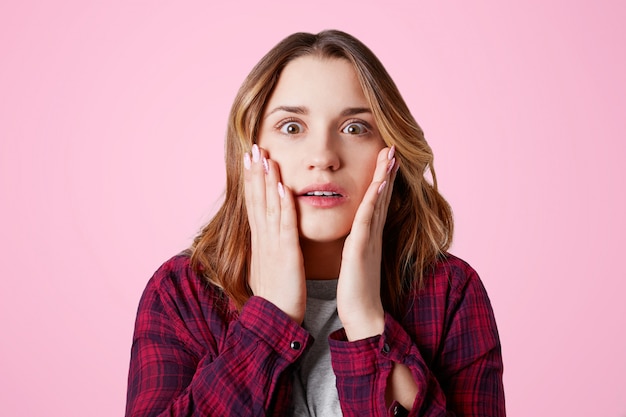 Stunned female model stares at camera, keeps hands on cheeks, being shocked with unexpected bad news