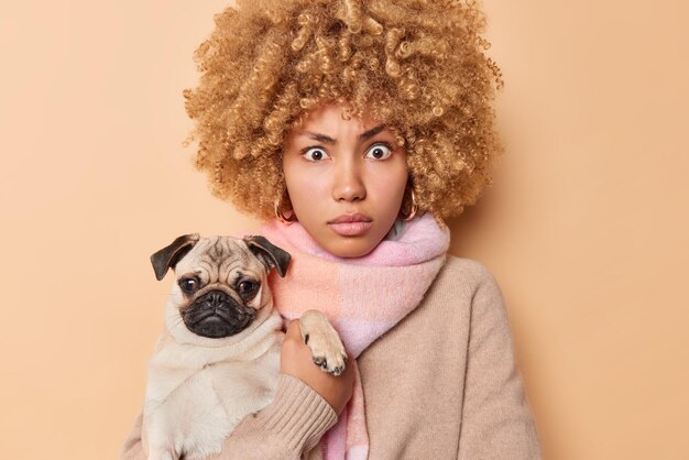 Stunned female model with curly hair feels surprised carries pug dog hugs favorite pet make memorable shot wears scarf around neck isolated over brown background. People and animals concept.