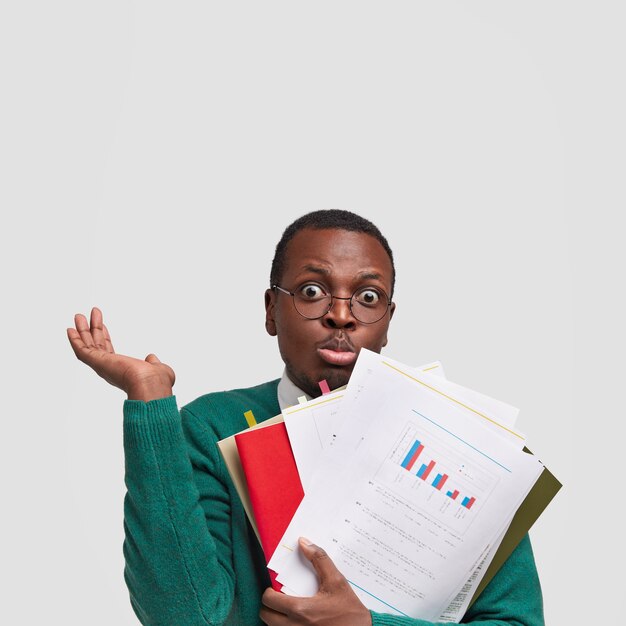 Stunned hesitant dark skinned guy shruggs shoulders and holds papers, textbooks, feels uncertain