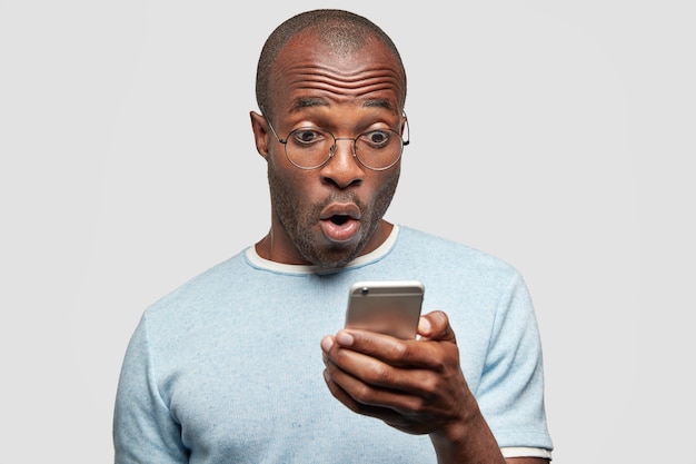 ошеломленный мужчина читает текстовое сообщение с удивленным выражением лица, держит сотовый телефон, узнает что-то шокирующее