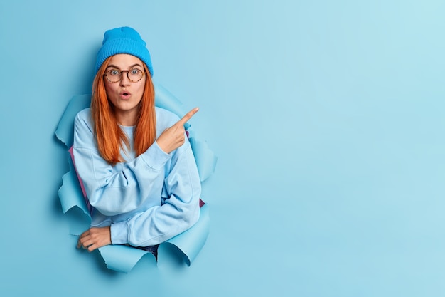 Free photo stunned redhead young woman points at upper right corner demonstrates copy space with surprised expression dressed in fashionable youth clothes.