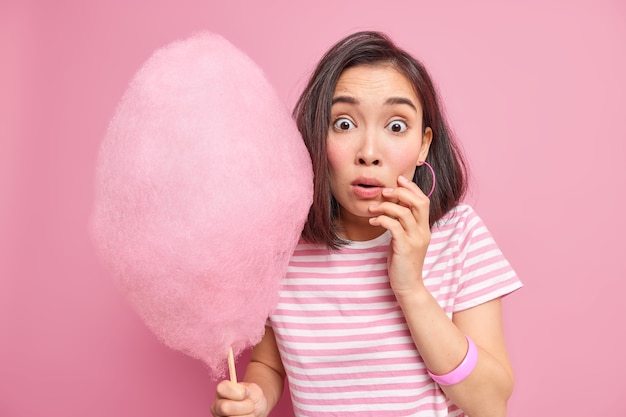 Бесплатное фото Ошеломленная испуганная брюнетка держит сладкую сахарную вату, удивленная услышать, что что-то удивительное узнает, сколько калорий она собирается съесть, носит полосатую футболку, изолированную над розовой стеной.