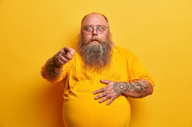 Free photo stunned shocked bearded obese man points index finger and holds tummy, reacts to overwhelming unexpected news, wears spectacles and yellow t shirt, poses indoor, feels impressed, excited