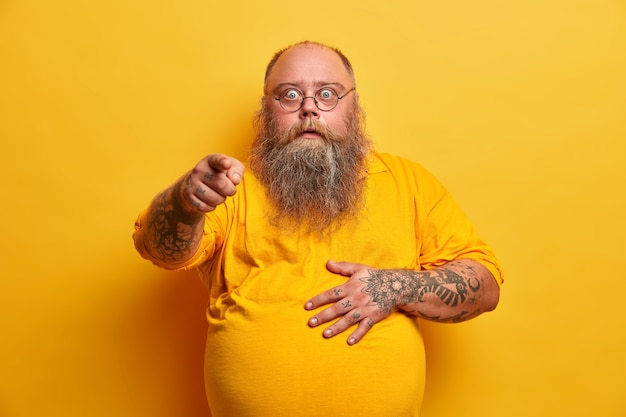 Ошеломленный шокированный бородатый тучный мужчина показывает указательным пальцем и держит живот, реагирует на неожиданные новости, носит очки и желтую футболку, позирует в помещении, чувствует себя впечатленным, взволнованным