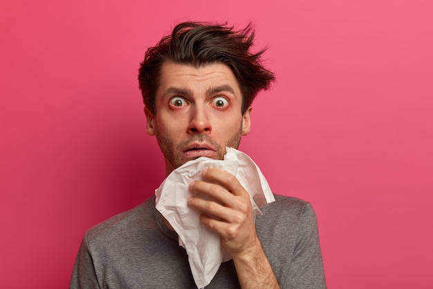 Stunned sick man has flu, virus or allergy respiratory, red watery eyes, blows nose in tissue, finds out about serious disease, poses over pink wall. Health, medicine and symptoms concept