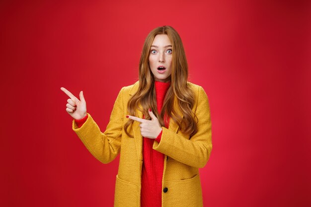 Stunned speechless and impressed attractive redhead woman with freckles in yellow coat drop jaw amazed as popping eyes at camera questioned, pointing left astonished over red background.
