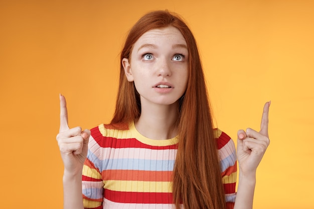 Stunned thrilled young redhead woman peer focused pointing up index fingers upwards look concentrated excited hold breath amused performance standing orange background intrigued and curious.