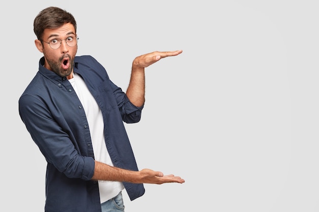 Stunned unshaven male with shocked facial expression gestures with hands, shows size or height of something, dressed in fashionable shirt, isolated over white wall, copyspace
