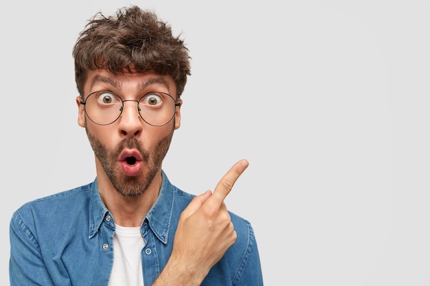 Stunned unshaven young man with wide opened eyes, points at upper right corner, being intrigued and suprirsed, poses against white wall. People and advertisement concept