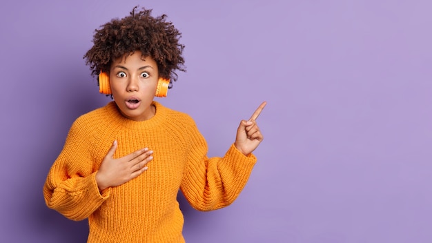 Stunned woman looks with bugged eyes and jaw dropped cannot believe in something astonishing wears orange sweater and headphones points at upper right corner 