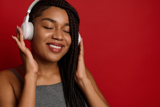 見事な美しいアフリカの女性は、コピースペースのある赤い背景に対して、ワイヤレスヘッドフォンで音楽を聴きながら、楽しさと喜びから目を閉じてポーズをとって、歯を見せる笑顔を浮かべます