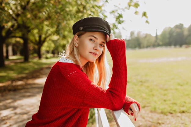 Stunning blonde enjoying sunny weather outdoor. Pretty girl in trendy red pullover looking good in the autumn park.