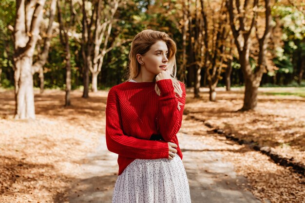 Stunning blonde in stylish clothes looking far in the autumn forest. Lovely girl enjoys good weather outdoor.