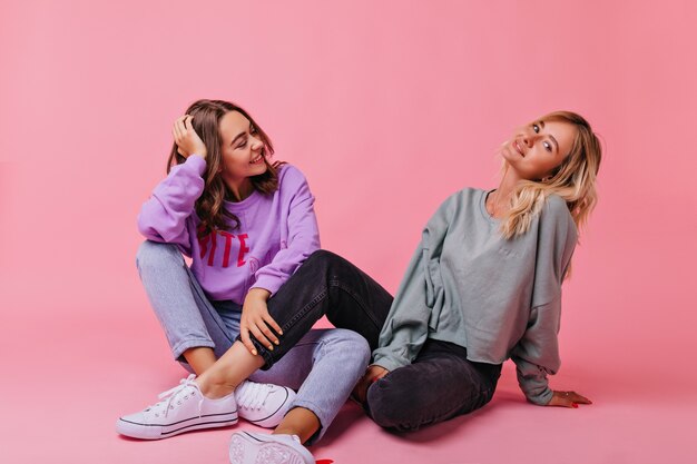 Stunning blonde woman in black pants and white shoes sitting on pink. Relaxed cute sisters posing on the floor.