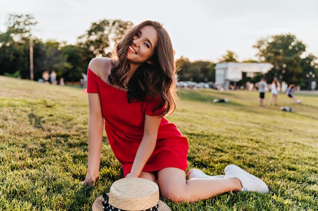 Сногсшибательная брюнетка женская модель сидит на земле с веселой улыбкой. Открытый выстрел возбужденной девушки в красном платье позирует на траве.