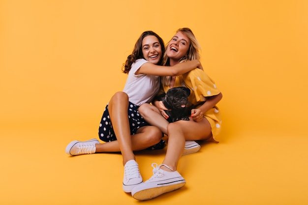 Stunning brunette girl in white shoes embracing her sister with happy smile. Carefree blonde lady having fun with best friend and bulldog during portraitshoot on yellow.