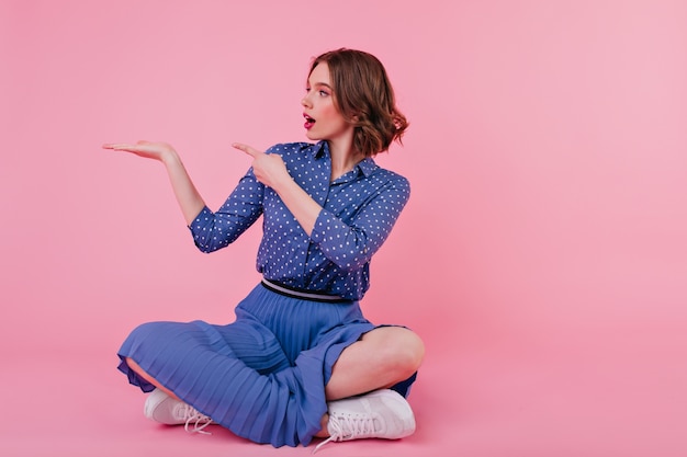 Stunning caucasian girl in white sneakers sitting with legs crossed. Adorable young woman in blue blouse posing on the floor on pink wall.