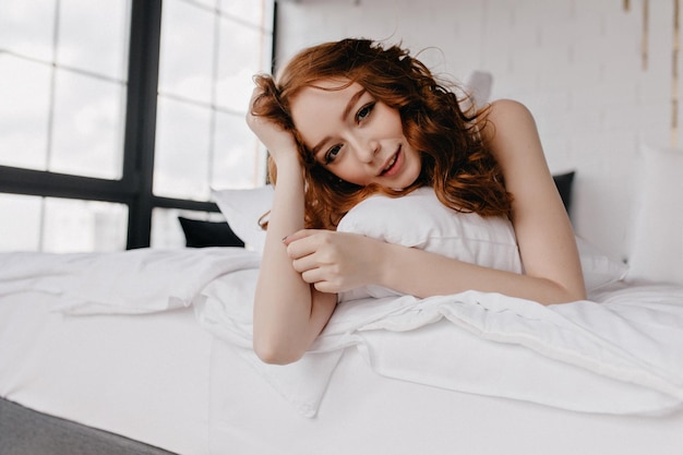 장난스런 미소로 카메라를 바라보는 물결 모양의 머리를 가진 멋진 백인 소녀 침대에서 쉬고 있는 아름다운 백인 여성의 실내 사진