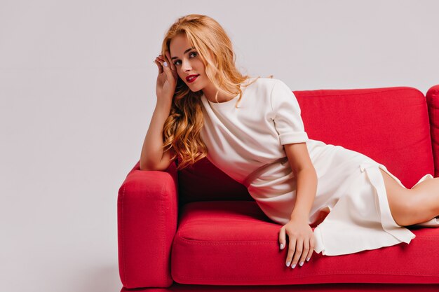 Stunning caucasian woman in white dress lying in living room. Indoor shot of graceful female model with wavy hair chilling on couch.