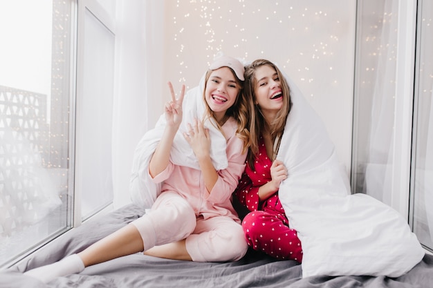 Stunning curly woman in sleepmask expressing happiness while posing in bedroom. Two european girls in pajamas sitting under blanket.