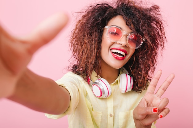 Stunning curly woman with bronze skin posing with peace sign. pleased black girl in yellow shirt and big white earphones.