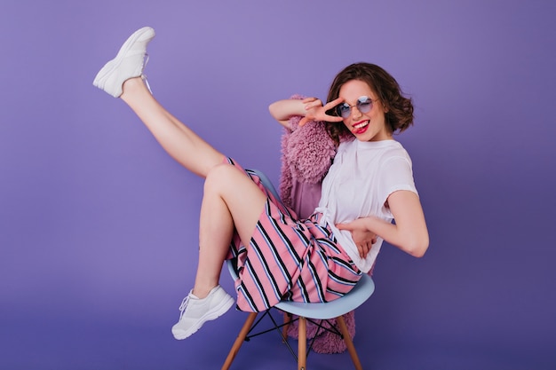 Сногсшибательная девушка в солнечных очках позирует на фиолетовой стене. Брюнетка жизнерадостная молодая женщина в белых туфлях, сидя на стуле с улыбкой.