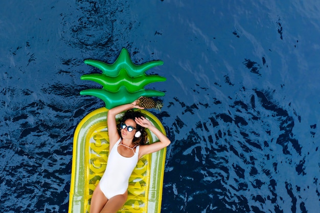 Foto gratuita splendida donna europea in costume da bagno sdraiato sul materasso di ananas. adorabile ragazza sottile in occhiali da sole rilassarsi in piscina nella mattina d'estate.