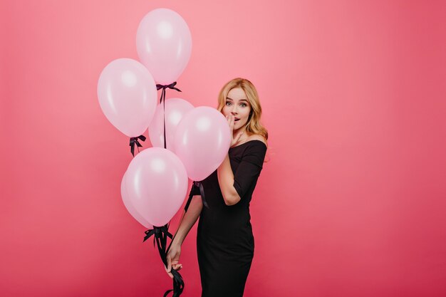 Потрясающая светловолосая женщина выражает удивленные эмоции, держа в руках букет воздушных шаров. Потрясенная молодая стройная дама в черном платье, стоя на розовой стене.