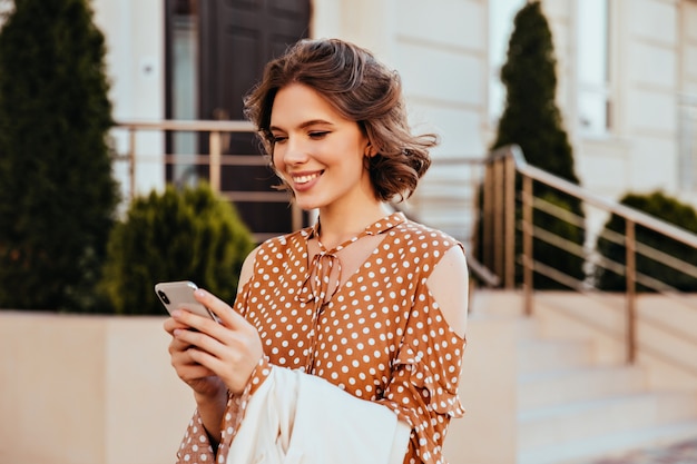 Stunning female model in elegant blouse looking at phone screen with interested face expression. Outdoor shot of pleased european woman in brown attire texting message with smile.