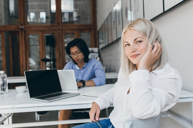 Stunning female programmer playing with blonde hair while posing at workplace with asian male colleague. Busy chinese web-developer working with laptop sitting at the table with white secretary girl.