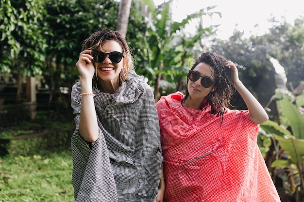 Stunning female tourists with wet hair posing after rain. Outdoor portrait of smiling travelers in sunglasses and raincoats standing on nature.