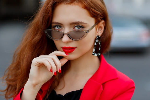 Stunning ginger female looking forward, holding glasses. Dressed in casual red jacket.