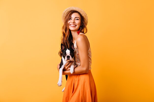 프랑스 불독을 들고 여름 옷에 멋진 생강 소녀. 강아지와 함께 초상화 촬영하는 동안 웃 고 모자에 매력적인 젊은 아가씨.