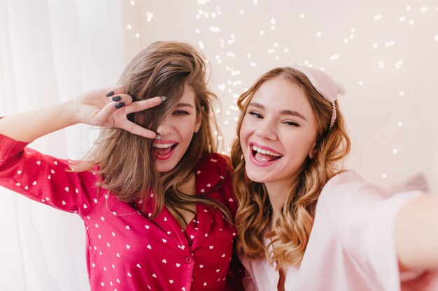 Stunning girl in red night-suit posing with peace sign near sister. Adorable curly lady in eyemask making selfie with her female friend.