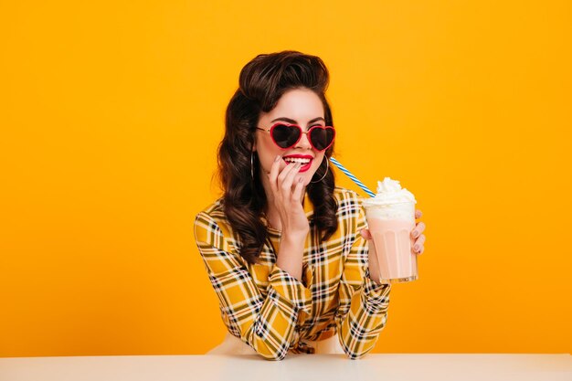 ミルクシェイクを飲むサングラスの見事な女性黄色の背景でカクテルを楽しんで笑っているピンナップガールのスタジオショット