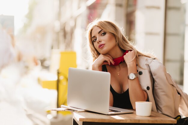 Stunning lady wears necklace and wristwatch looks to camera during work with laptop in cafe