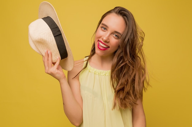 Потрясающая длинноволосая женщина-модель позирует на желтой стене, держит шляпу и улыбается