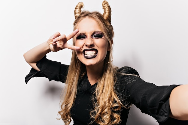 Foto gratuita incredibile strega dai capelli lunghi che scherza ad halloween. tiro al coperto di signora bionda che fa selfie in costume malvagio.