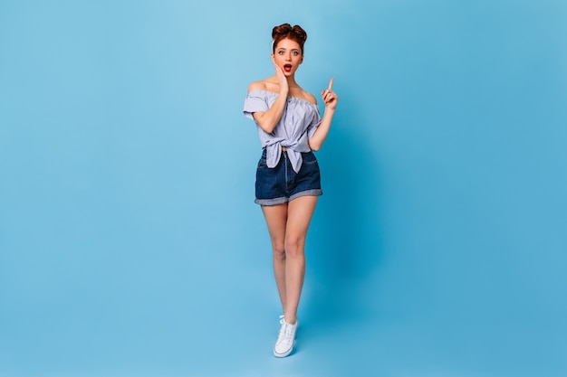 Stunning pinup woman pointing up with finger. Full length view of surprised ginger lady wears denim shorts.