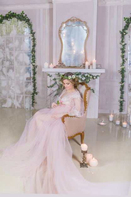 Stunning pregnant woman in pink dress rests on couch surrounded with shiny candles