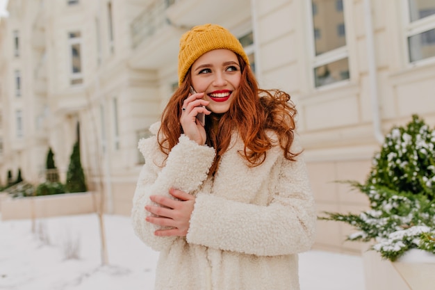 Потрясающая рыжая дама улыбается, позируя с телефоном. Открытый выстрел привлекательной имбирной женщины, стоящей на улице зимним утром.
