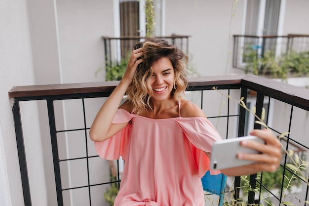 Stunning tanned woman playing with her wavy hair while posing at balcony. Laughing caucasian girl using smartphone for selfie and expressing positive emotions.